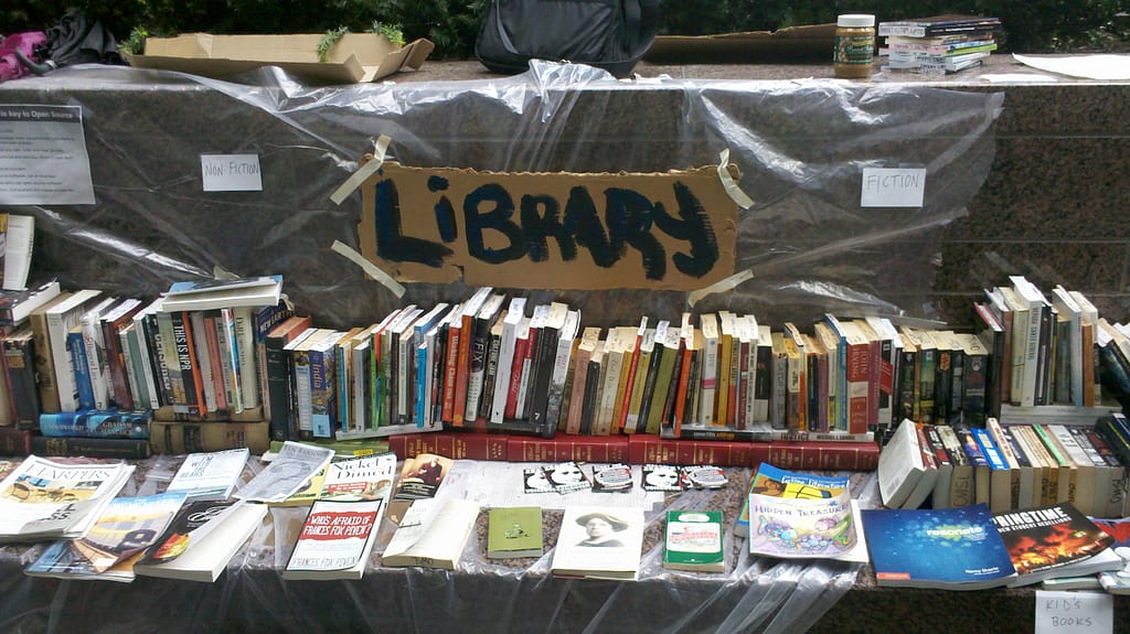 Occupy Wall Street Library image of books in Zuccotti Park, spread out on plastic sheeting with a placard announcing LIBRARY taped to the back wall.