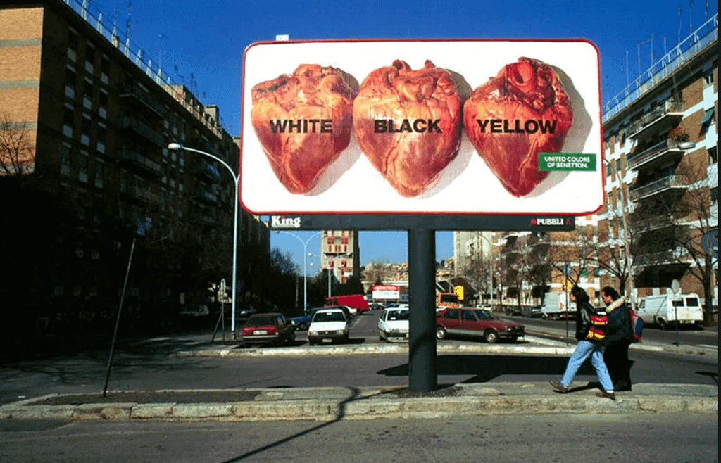 Two people walking under a billboard in an urban setting. Image on billboard is of three organs that appear to be human hearts side by side labelled "white" "black" "yellow" with the green United Colors of Benetton logo.